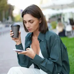 Kobieta z oliwkowym kolorem skóry, trzymająca kawę i telefon, siedzi na ławce w mieście.
