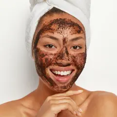 Uśmiechnięta kobieta z ręcznikiem na głowie ma na twarzy naturalny peeling z kawy.