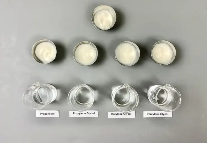 Sześć próbek kremów w małych pojemnikach i cztery zlewki z płynami: propanediol, glikol propylenowy, **butylene glycol** i pentylene glycol.