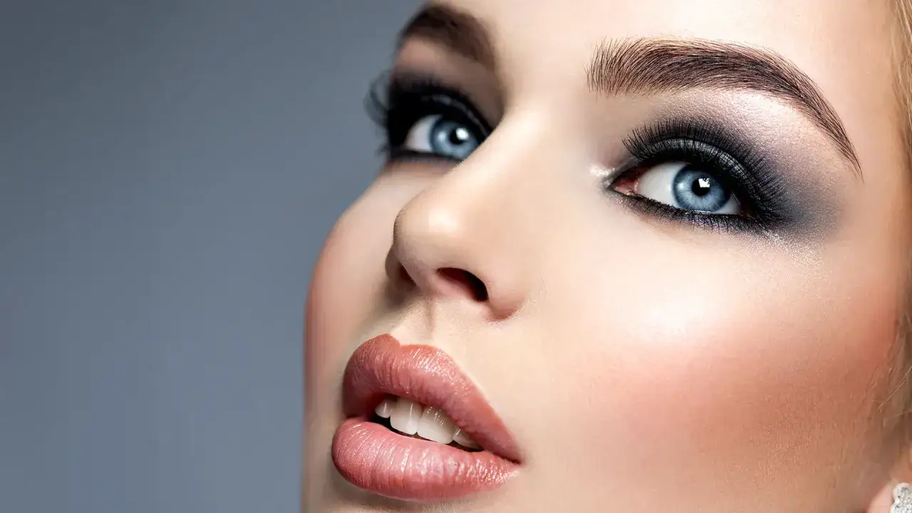 Niebieskie oczy podkreślone intensywnym makijażem smokey eyes, z błyszczącymi ustami i delikatnym rumieńcem na policzkach.
