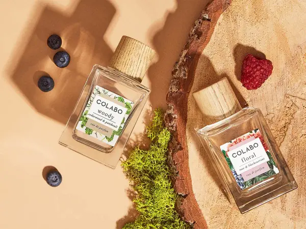 Dwa flakony perfum COLABO: "woody" z jagodami i "floral" z maliną, otoczone mchem i drewnem.